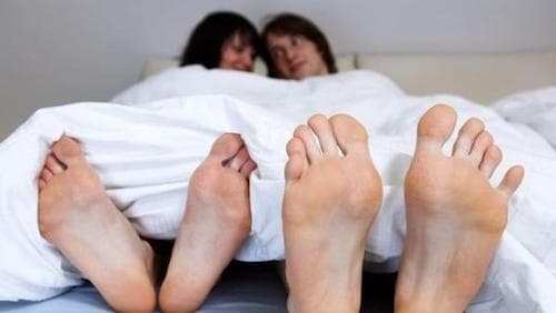 Im Fokus des Bildes stehen zwei Fußpaare die von einer Bettdecke bedeckt sind.